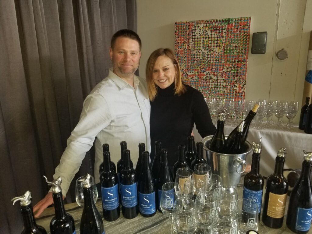 a man and a woman standing behind a table full of bottles of wine.